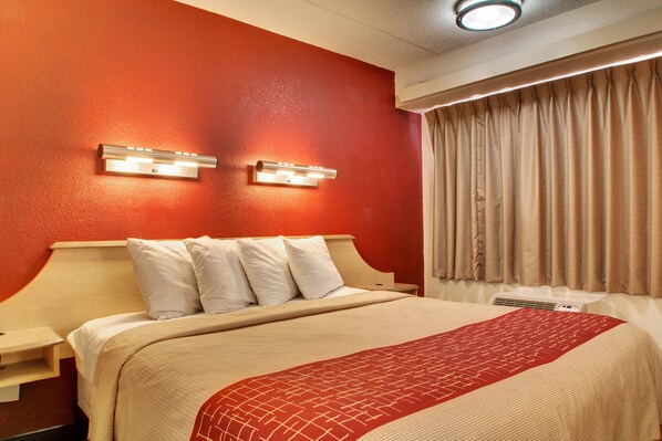 A hotel room with a large bed, two bedside tables, a wall-mounted light fixture, a window with curtains, and an air conditioning unit. - Red Roof Inn St Louis - Westport/ Maryland Heights (Maryland Heights)