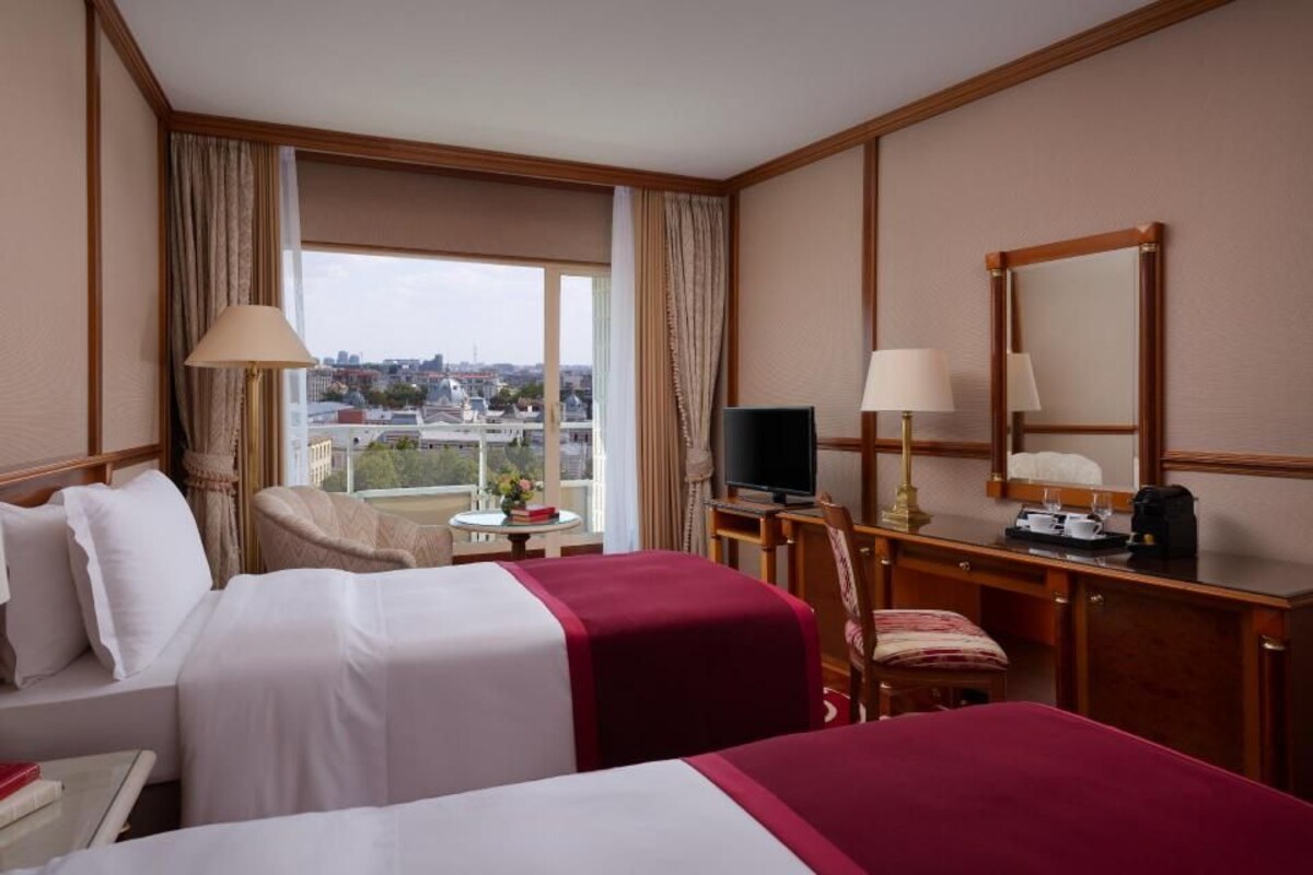 A hotel room with two beds, a desk with a television, a chair, and a view of the city.