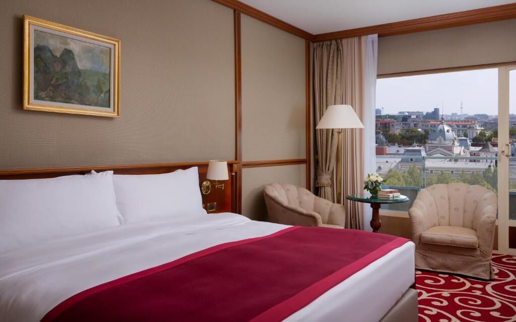 Ein Hotelzimmer mit einem großen Bett, einem Bild an der Wand, Blick auf die Stadt durch das Fenster und einem Sessel am Fenster.
