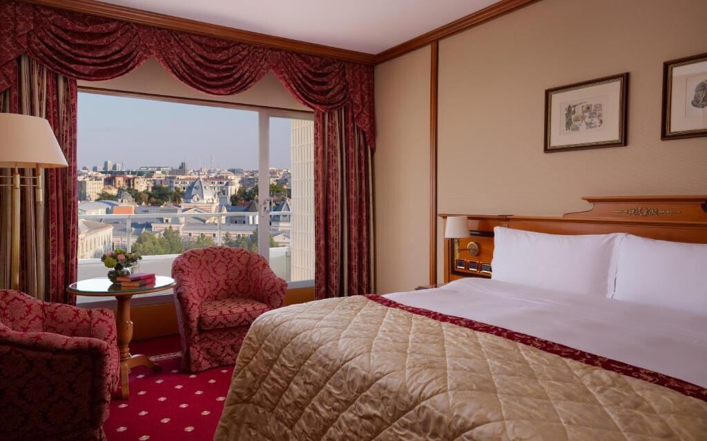 Ein Hotelzimmer mit einem großen Bett, einem Sessel, einem kleinen Tisch mit einer Vase und Blick auf die Stadt durch ein großes Fenster.
