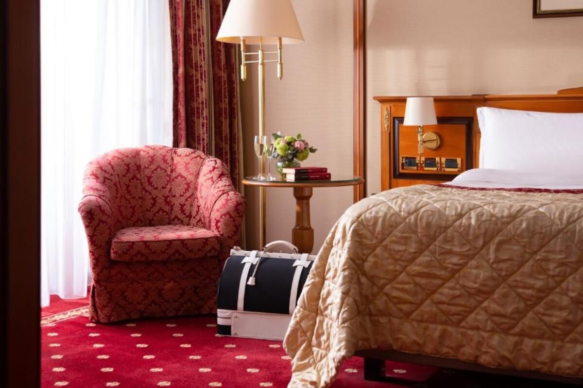 A hotel room with a bed, a chair, a table with a lamp, a clock, and a small vase with flowers.
