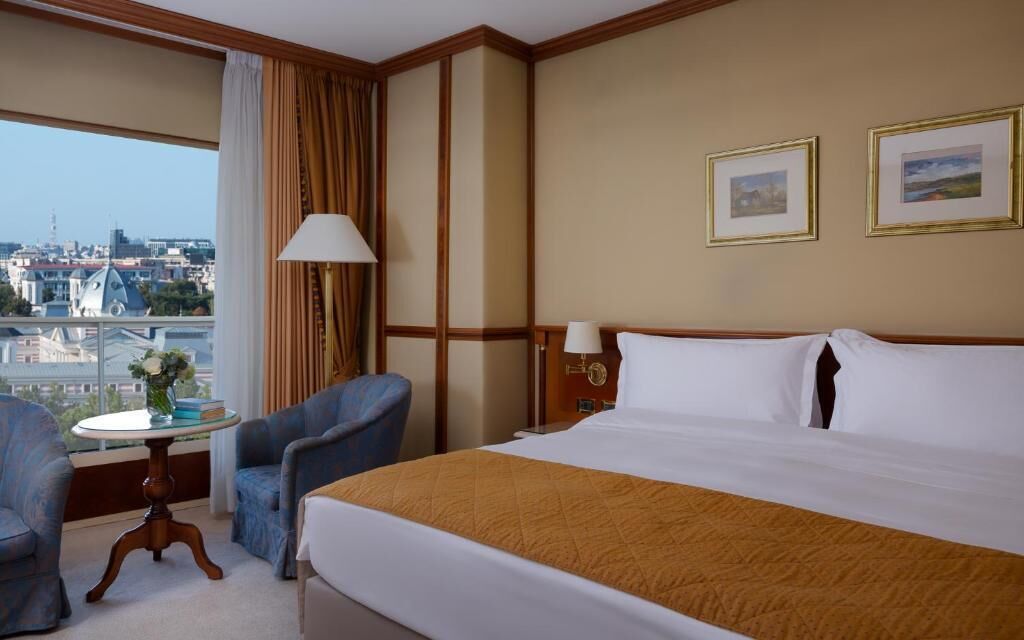 Ein Hotelzimmer mit einem großen Bett, zwei Sesseln, einem kleinen Tisch mit einer Vase, einer Lampe und gerahmten Bildern an der Wand.