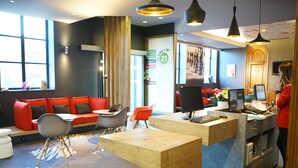 A modern reception area with a wooden desk, a red sofa, and a large window. - ibis Douai Centre (Douai)