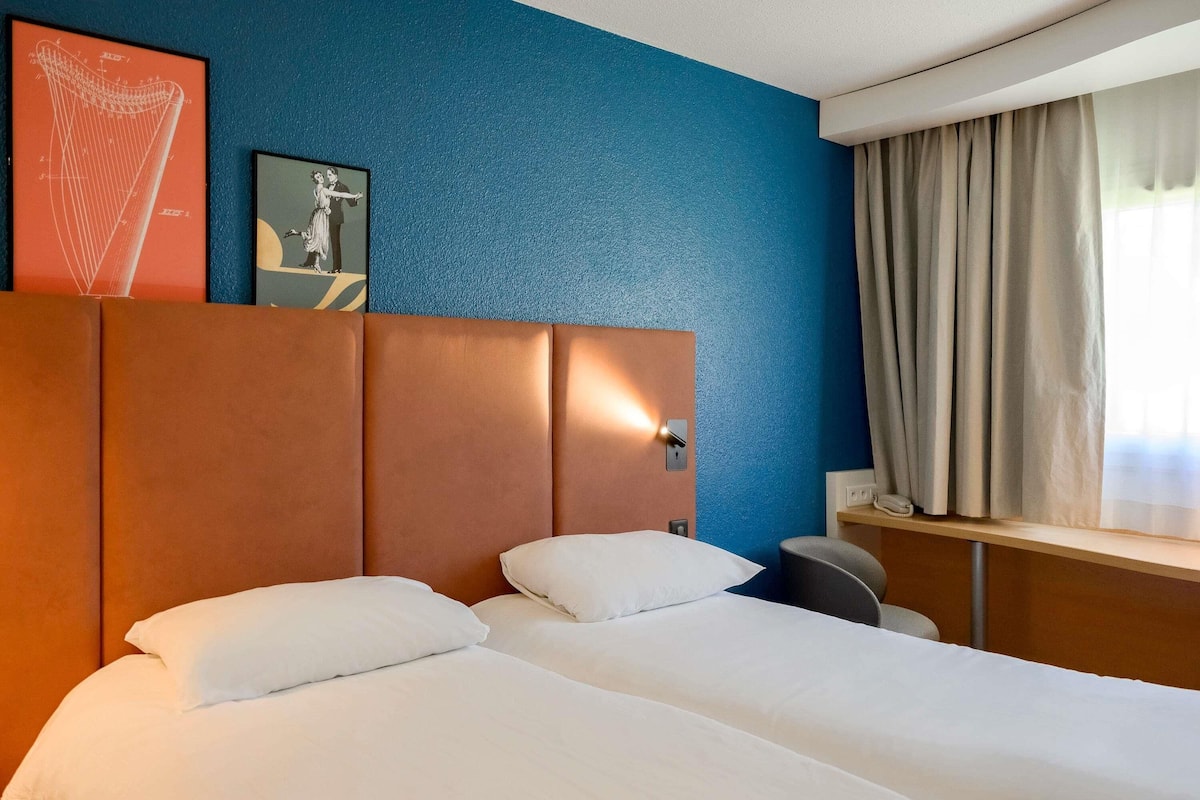 Habitación de hotel con cama, cabecera, un reloj de pared y una ventana con cortinas.