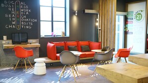 A modern lounge area with a red sofa, grey and red chairs, a wooden table, and a large window providing natural light. - ibis Douai Centre (Douai)