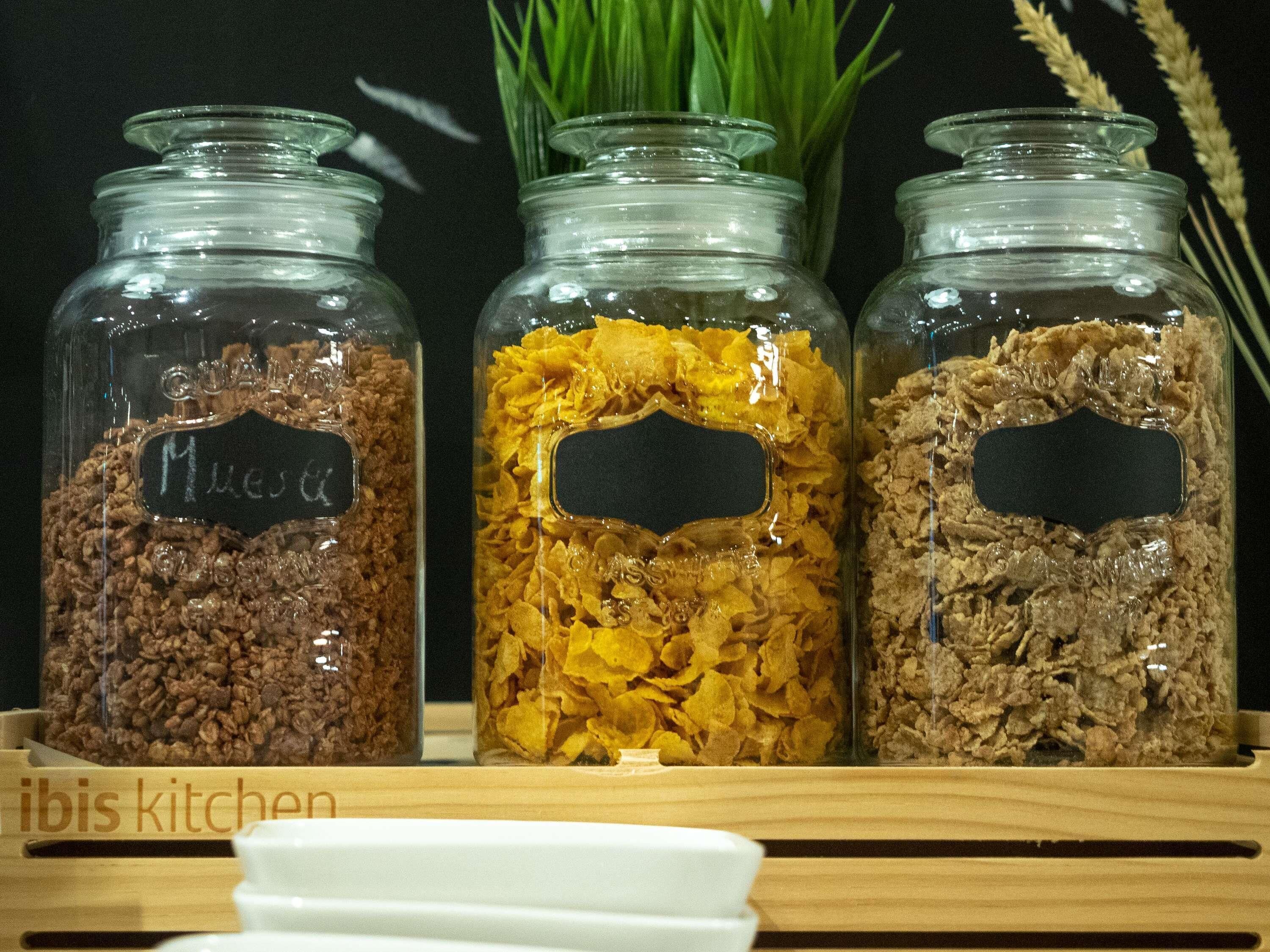 Tres frascos de vidrio con etiquetas, que contienen diferentes tipos de granos, exhibidos sobre una repisa de madera.