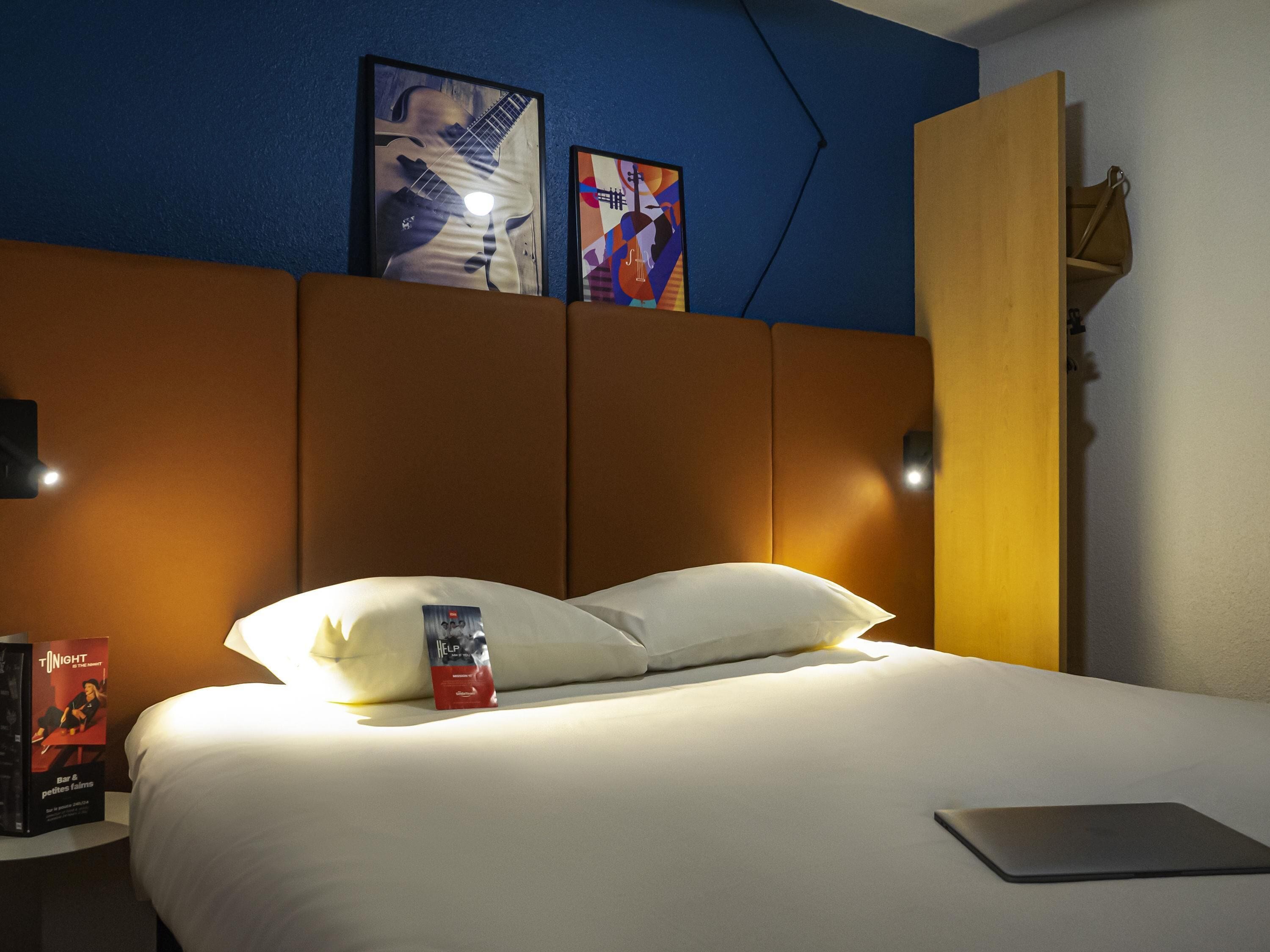Habitación de hotel con cama, almohadas, una computadora portátil, una mesita de noche con un libro y decoración en la pared.