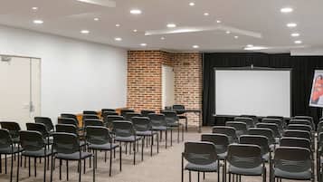 Un salón de conferencias con filas de sillas, una pantalla de proyector y una pared de ladrillo.