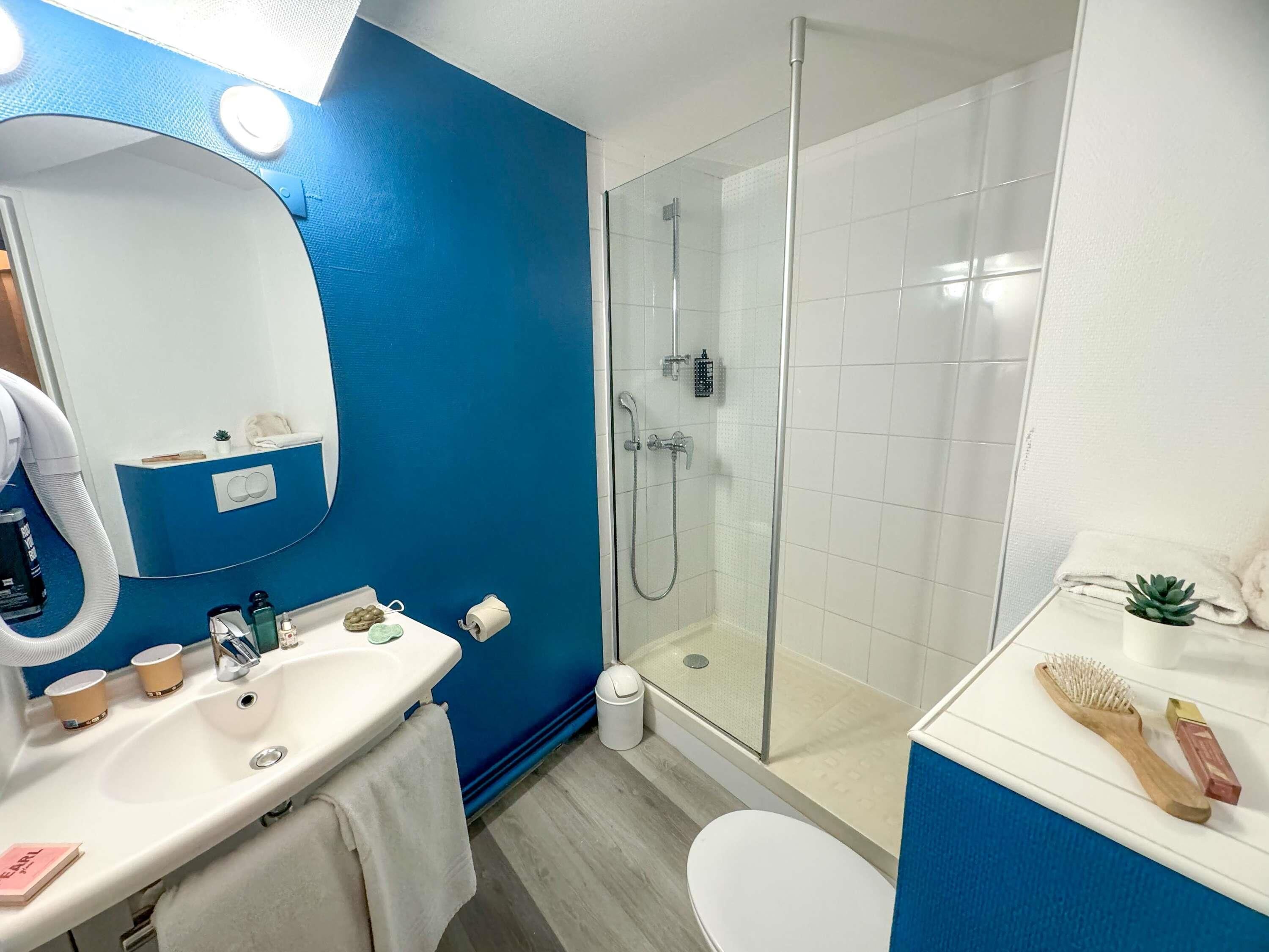 A modern bathroom with a blue accent wall, a glass-enclosed shower, a white sink with toiletries, and a small plant on the vanity.