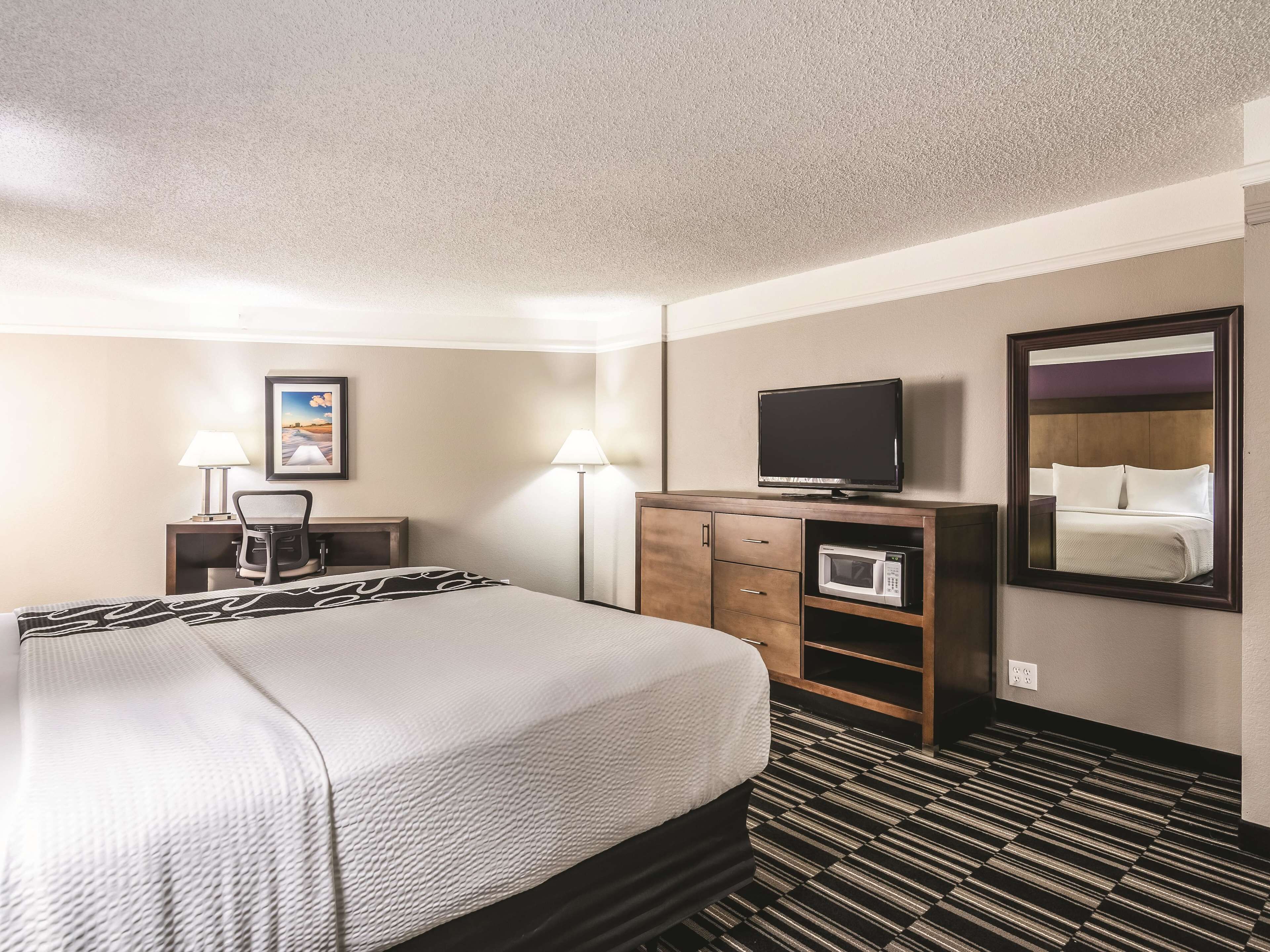 A hotel room with a bed, a desk, a chair, a television on a cabinet, a microwave, and a large mirror.