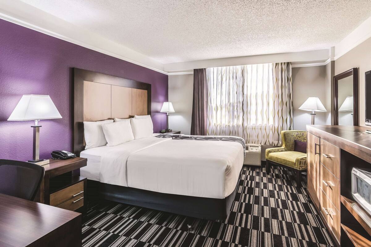 Una habitación de hotel con una cama grande, dos mesitas de noche con lámparas, un escritorio, una silla, un aparador y un amplio ventanal con cortinas.