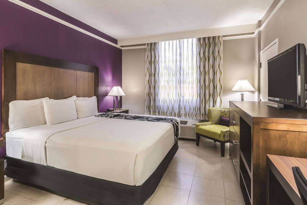 Una habitación de hotel con cama, dos mesitas de noche, televisor, una silla y ventana con cortinas.