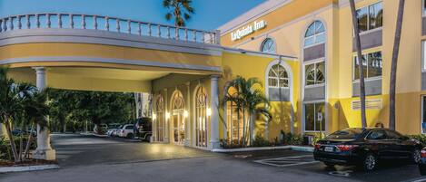 A hotel entrance with a curved balcony, palm trees, and a parking lot.