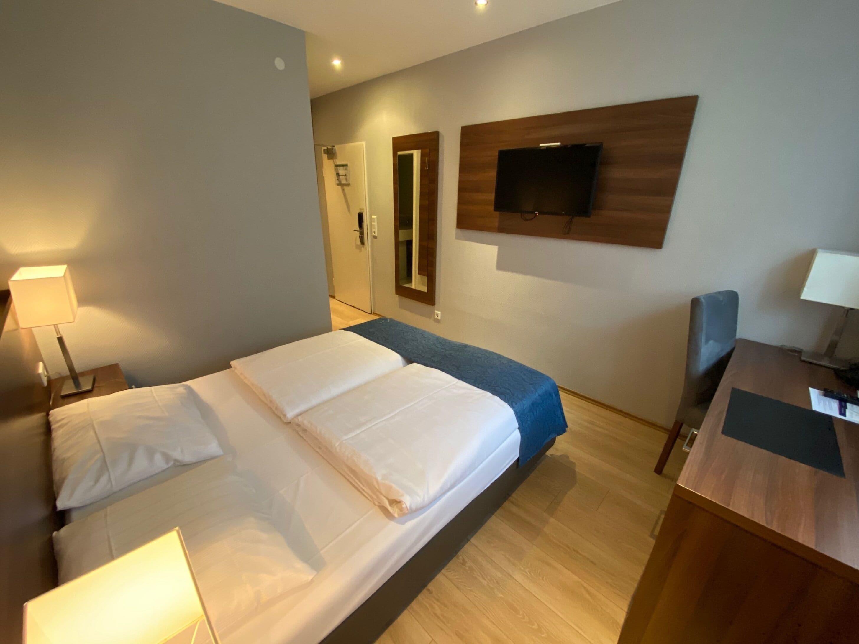 Ein Hotelzimmer mit Bett, Nachttisch, Lampe, Fernseher und Schreibtisch mit Stuhl.