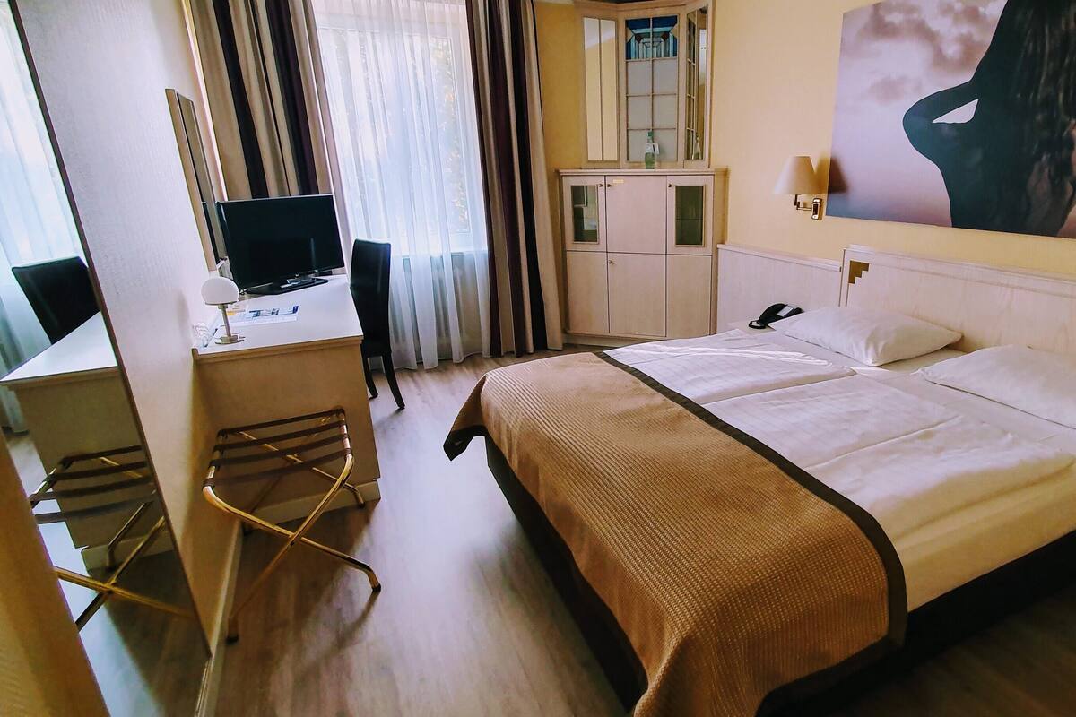 Quarto de hotel com cama, escrivaninha com computador, cadeira, guarda-roupa e uma janela ampla com cortinas.