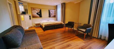 Habitación de hotel con cama, sofá, dos sillones, una mesita y ventana con cortinas.