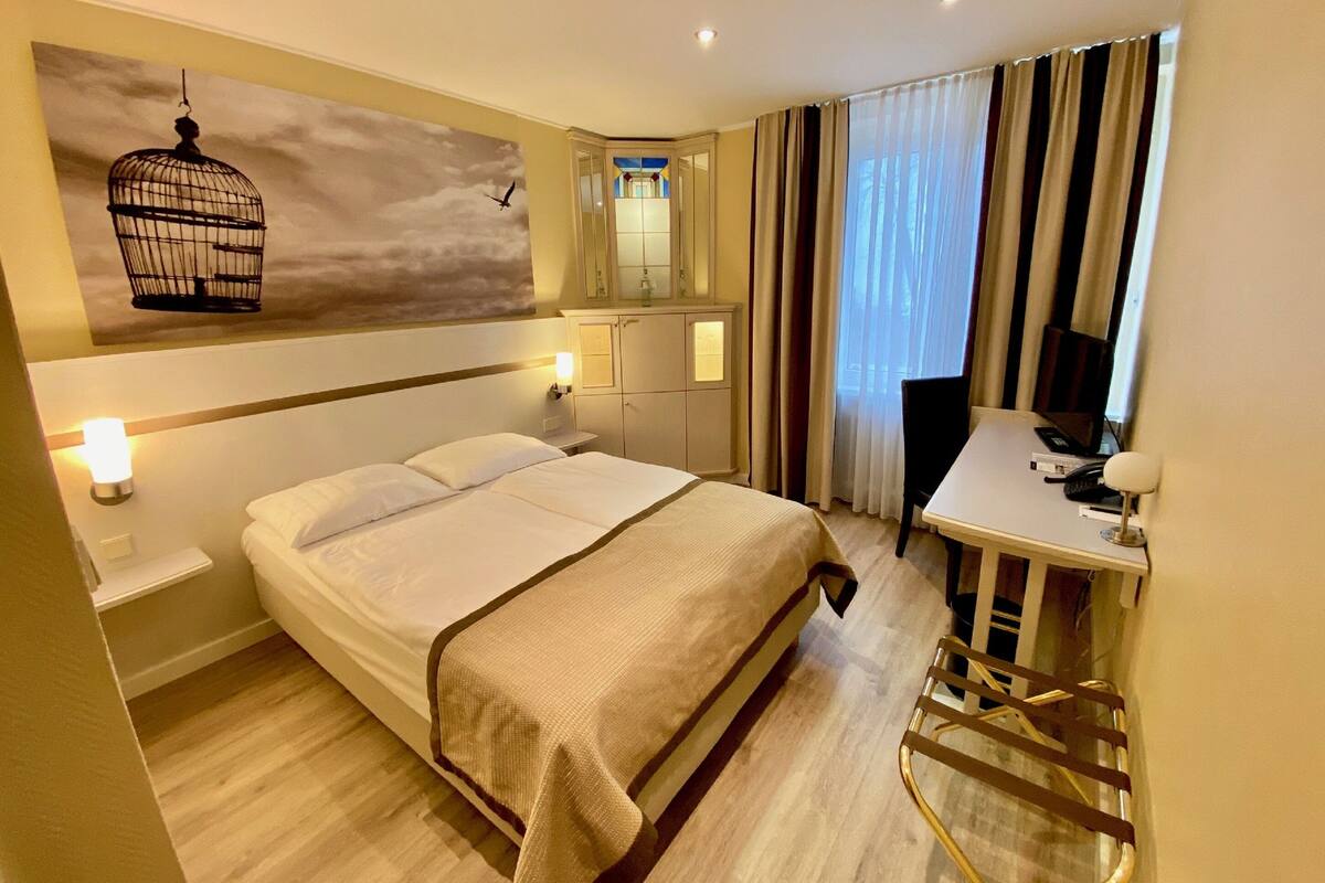 Quarto de hotel com uma cama, mesas de cabeceira, uma escrivaninha com cadeira, um espelho e uma janela com cortinas.