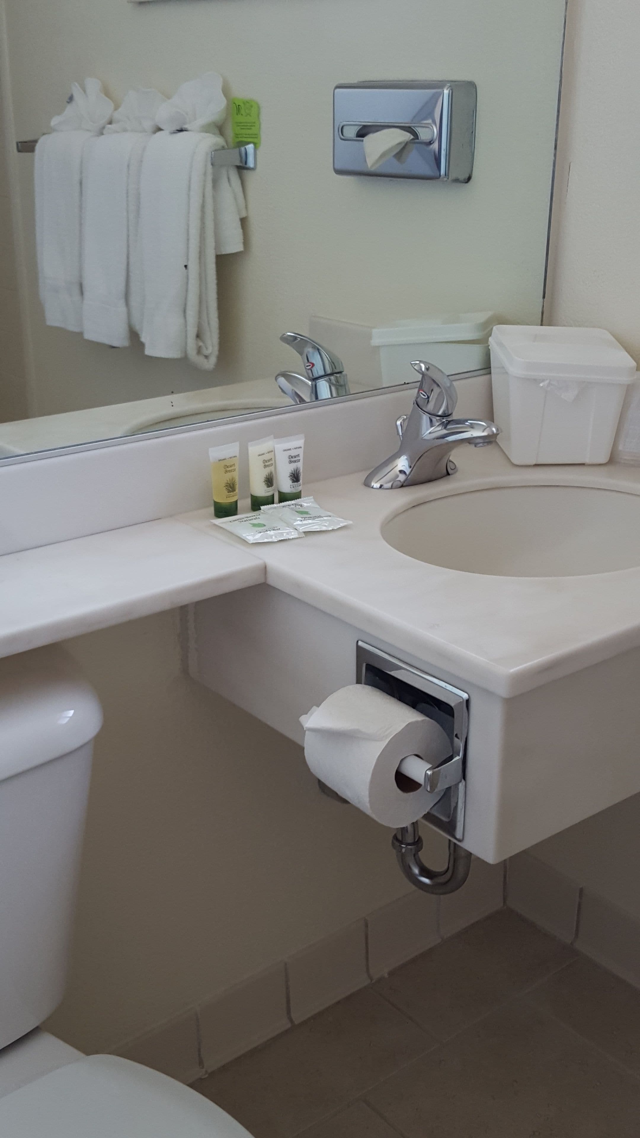 A bathroom with a white sink, a mirror, a towel rack with white towels, a toilet paper holder, and a trash bin.