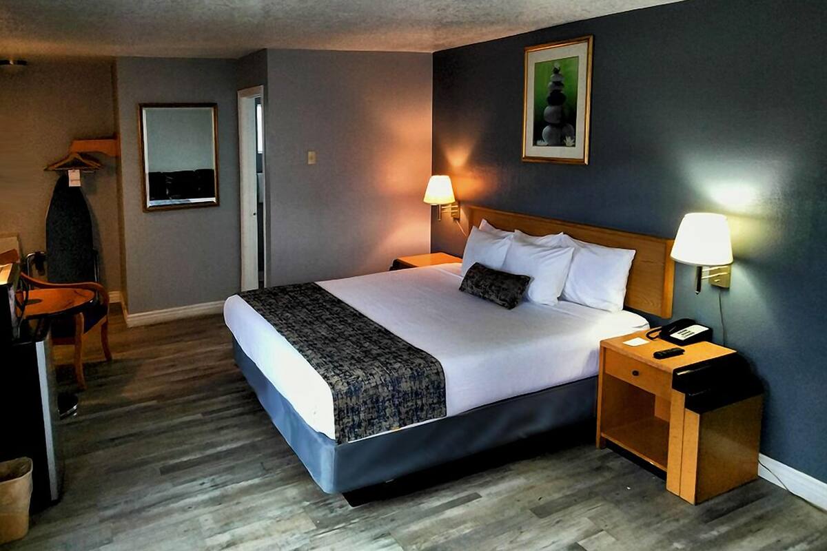Una habitación de hotel con una cama grande, dos lámparas de noche, una mesita de madera, una silla y un cuadro en la pared.