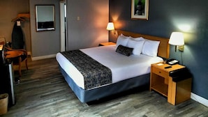 Hotellihuone, jossa on suuri sänky, kaksi yöpöytävalaisinta, puinen yöpöytä, tuoli ja seinällä oleva kehystetty kuva.