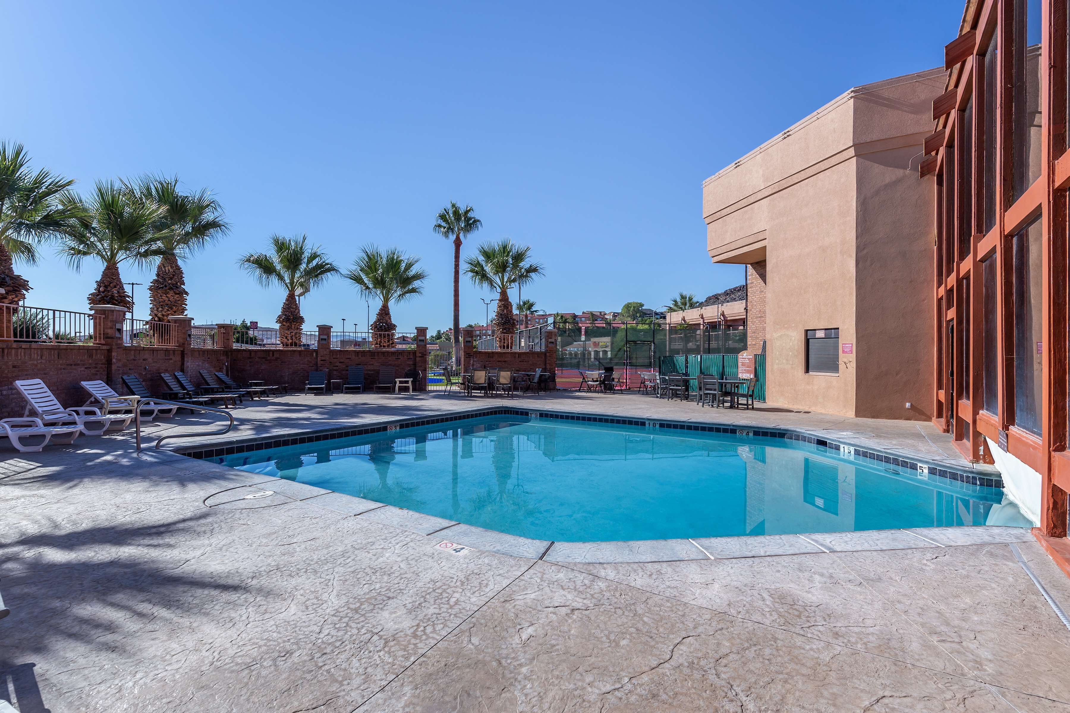 A swimming pool with lounge chairs and palm trees, surrounded by a brick wall and a building with large windows.