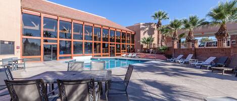 A pool area with lounge chairs and a table, a building with large windows, and palm trees.