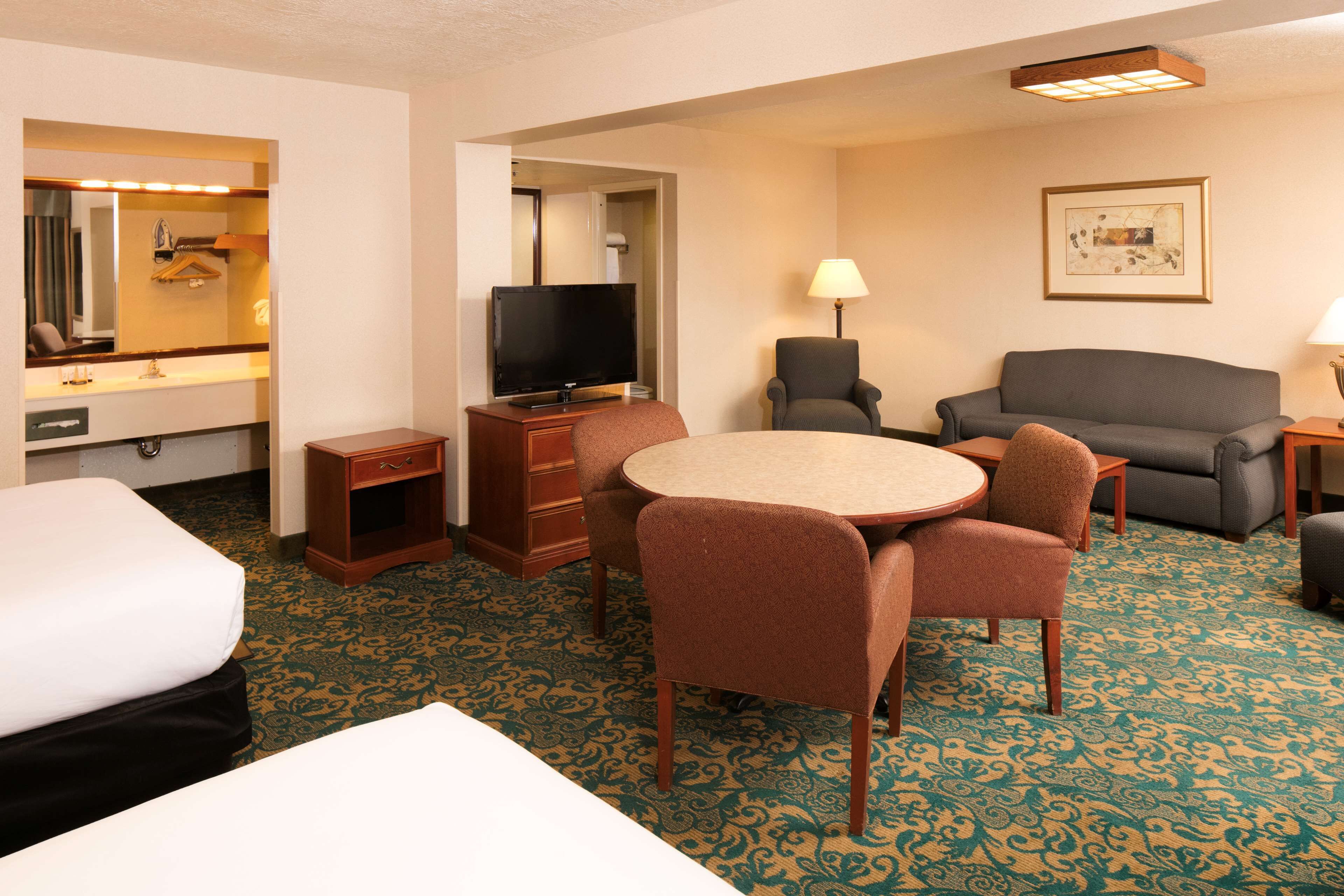 Habitación de hotel con cama, escritorio con televisor, mesa de comedor con sillas, sofá y alfombra estampada.
