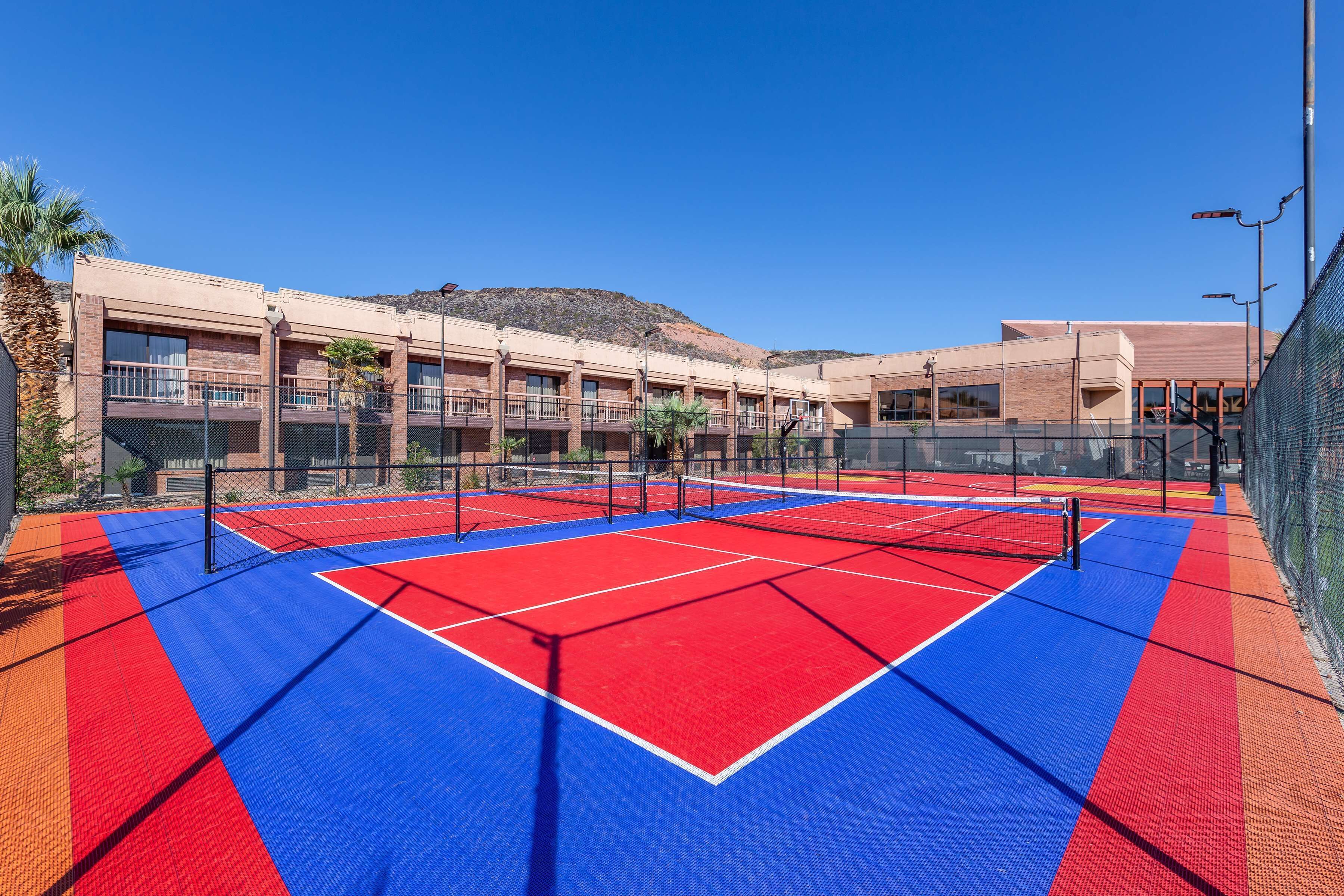 A tennis court with a red and blue surface, surrounded by a fence, and adjacent to a building with multiple windows and balconies.
