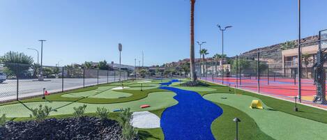 A golf course with a blue water feature, palm trees, and a fenced area with sports courts.