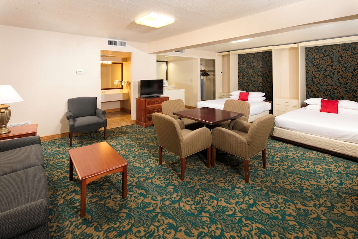 A hotel room with a sofa, two armchairs, a small table, a TV, a bed with red pillows, and a patterned carpet.