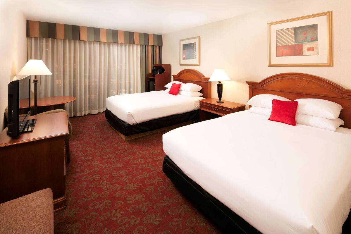A hotel room with two beds, a desk, a chair, a lamp, and a large window with curtains.