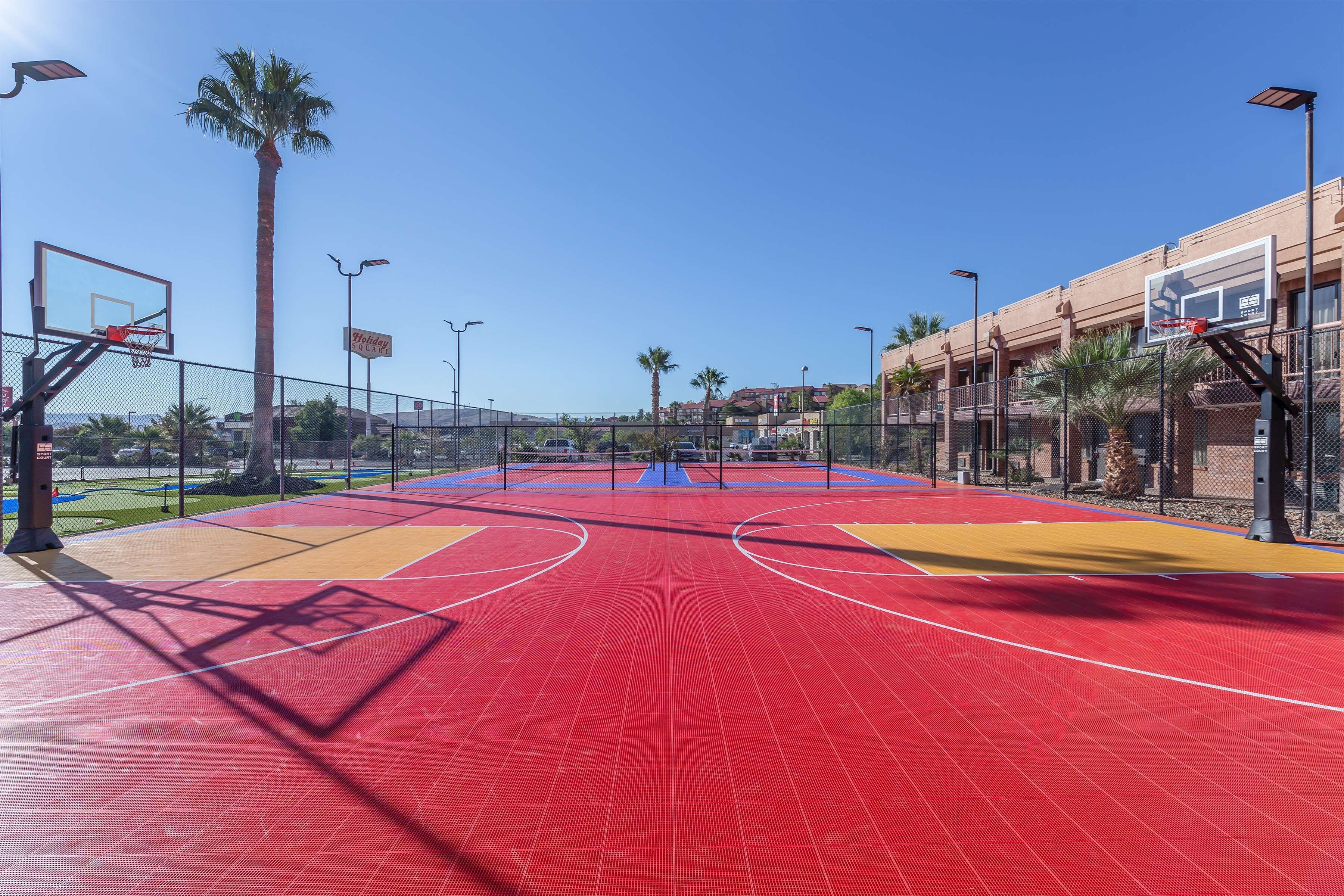 Una cancha de baloncesto al aire libre con superficie roja, rodeada por una cerca, palmeras y un edificio al fondo.