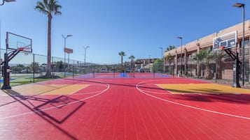 Una cancha de baloncesto al aire libre con superficie roja, rodeada por una cerca, palmeras y un edificio al fondo.