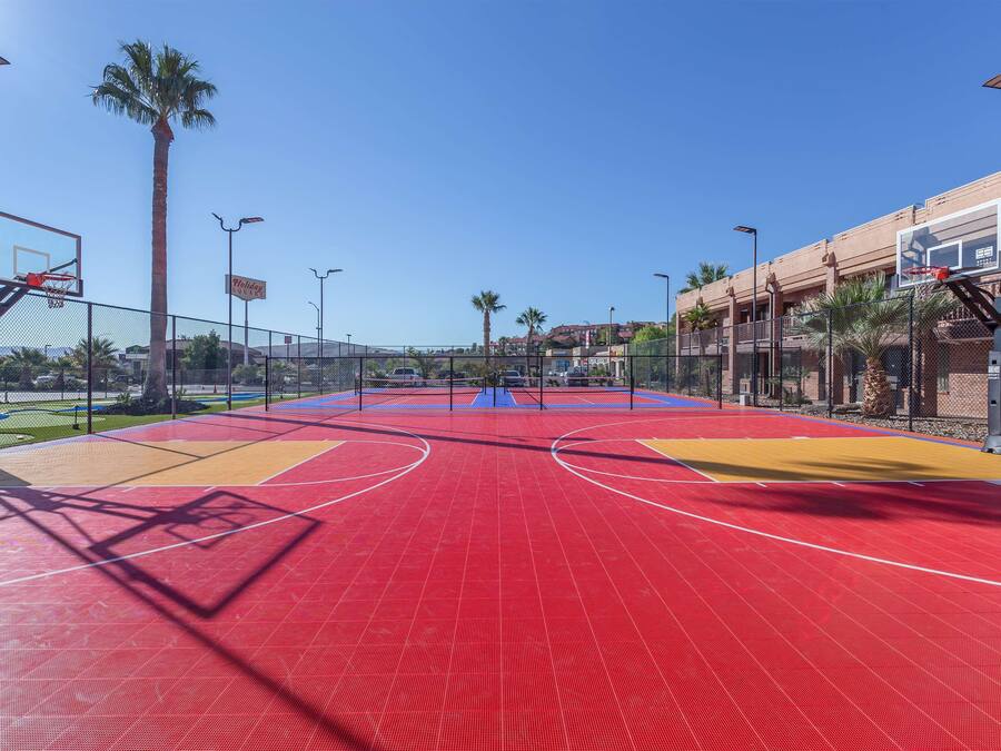 Una cancha de baloncesto al aire libre con superficie roja, rodeada por una cerca, palmeras y un edificio al fondo.