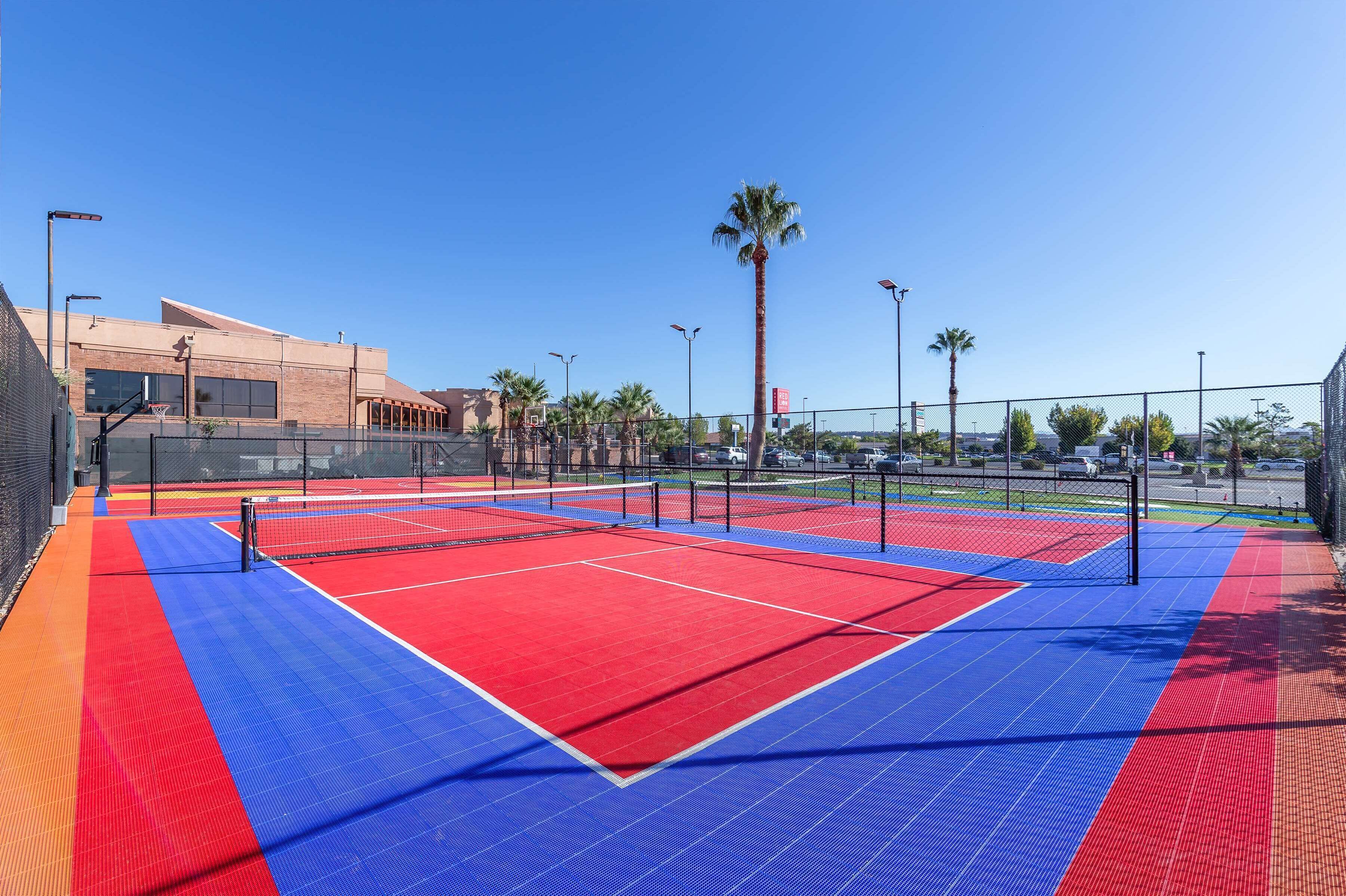 An outdoor tennis court with a red and blue playing surface, surrounded by a fence and palm trees.