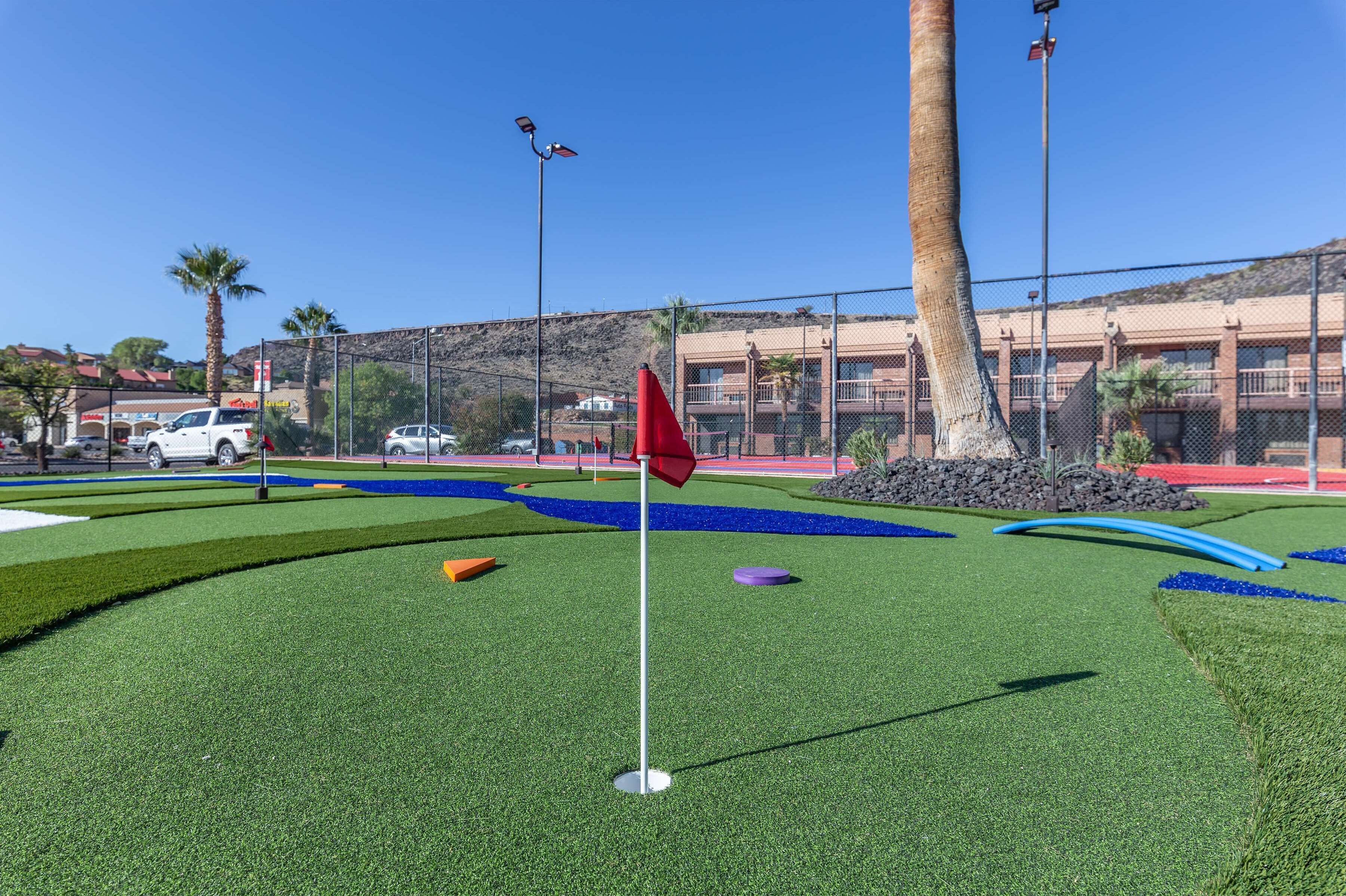 Un campo de mini golf con una bandera roja, palmeras y un edificio al fondo.