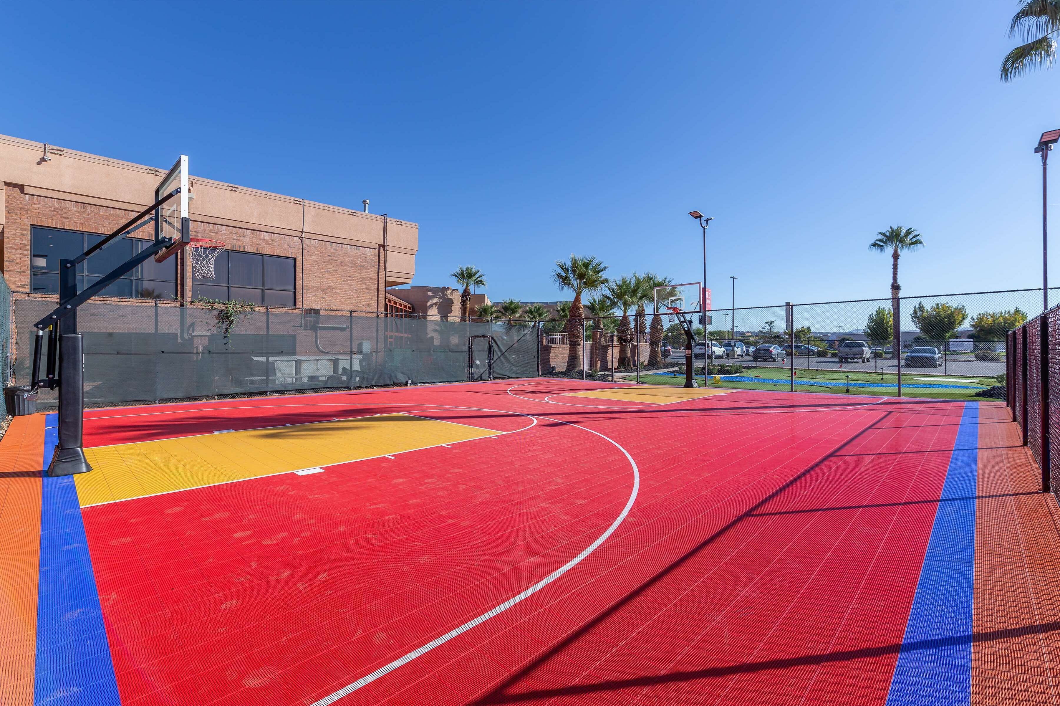 An outdoor basketball court with a red and yellow playing surface, surrounded by a fence and palm trees.