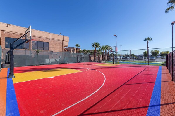An outdoor basketball court with a red and yellow playing surface, surrounded by a fence and palm trees.