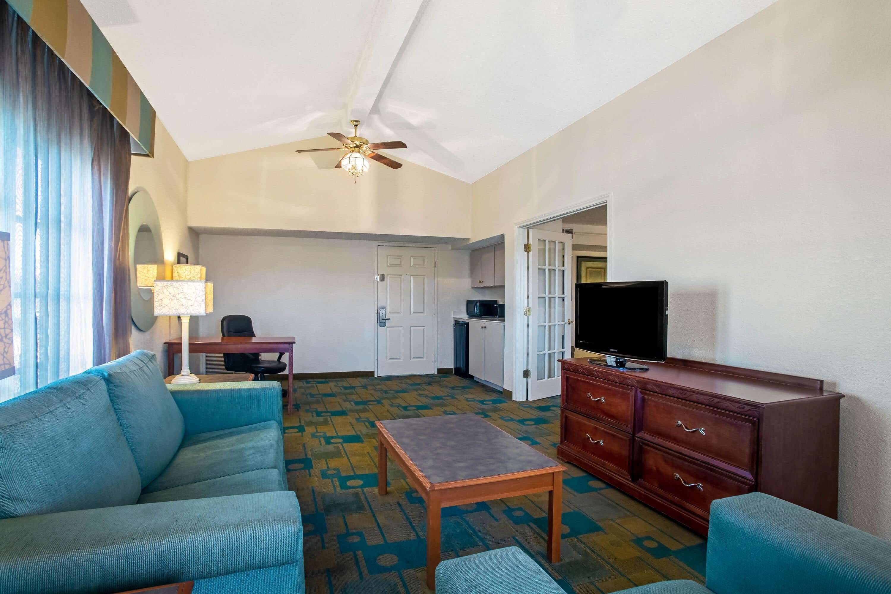 Une chambre d’hôtel avec un canapé bleu, une table en bois, un téléviseur, un ventilateur de plafond et un coin cuisine.