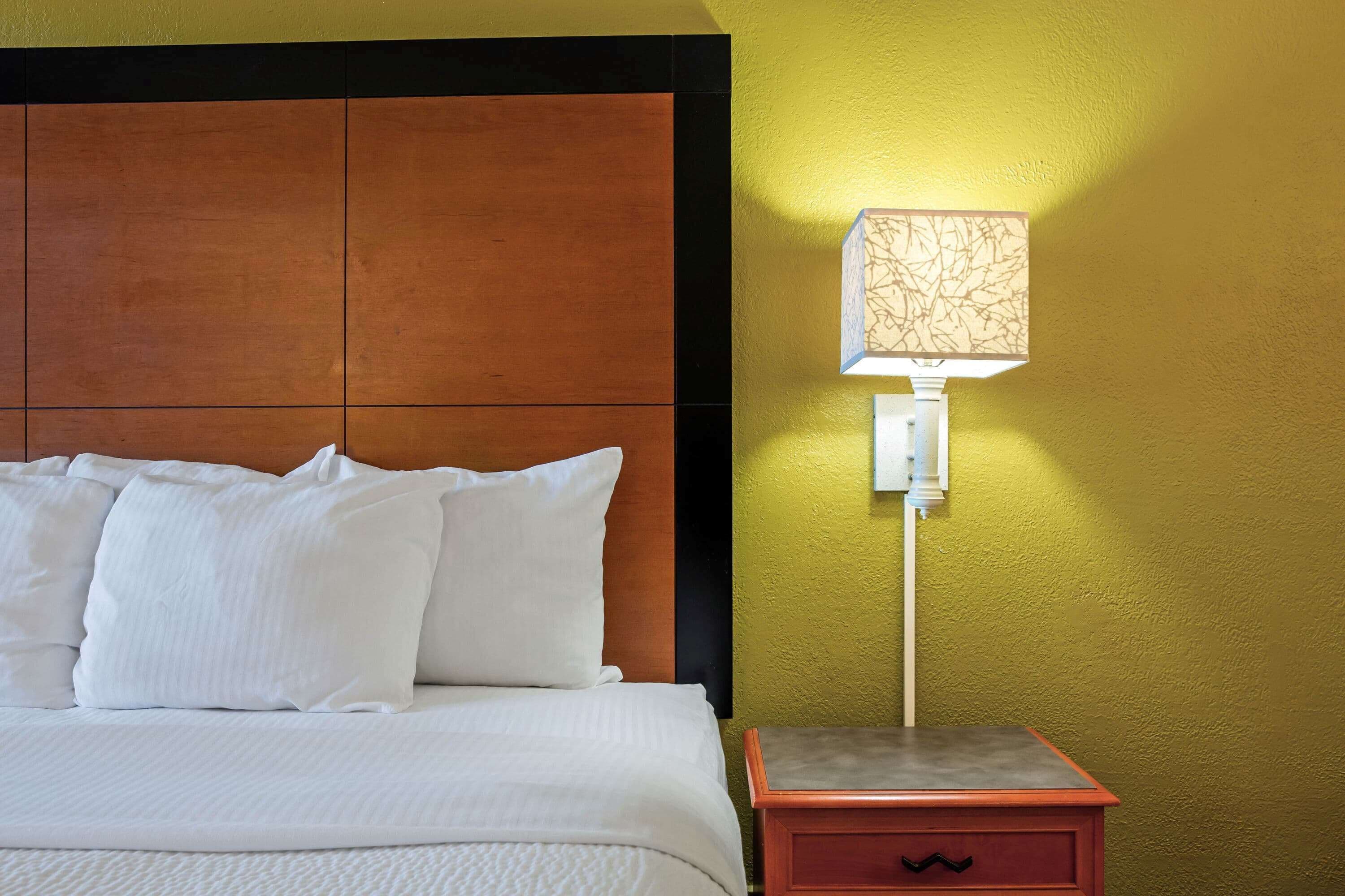 A neatly made bed with white linens and pillows, a wooden headboard, and a bedside table with a lamp against a yellow wall.