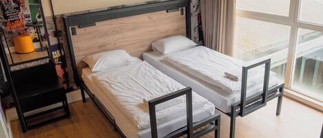 A room with two beds, a wooden floor, a window with blinds, and a mirror.