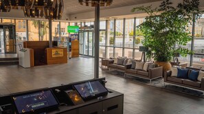 A modern hotel lobby with a reception desk, self-check-in kiosks, seating area, and large windows offering views of the city.