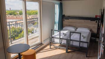 Un dormitorio con un ventanal grande, una cama con ropa de cama blanca, una mesita redonda pequeña y un cabecero de madera.