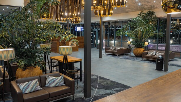 A spacious lobby with modern furniture, large potted plants, and a high ceiling with hanging lights.