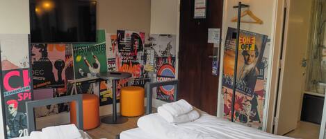 Habitación de hotel con dos camas, un televisor, carteles en la pared y una puerta que da a un baño.