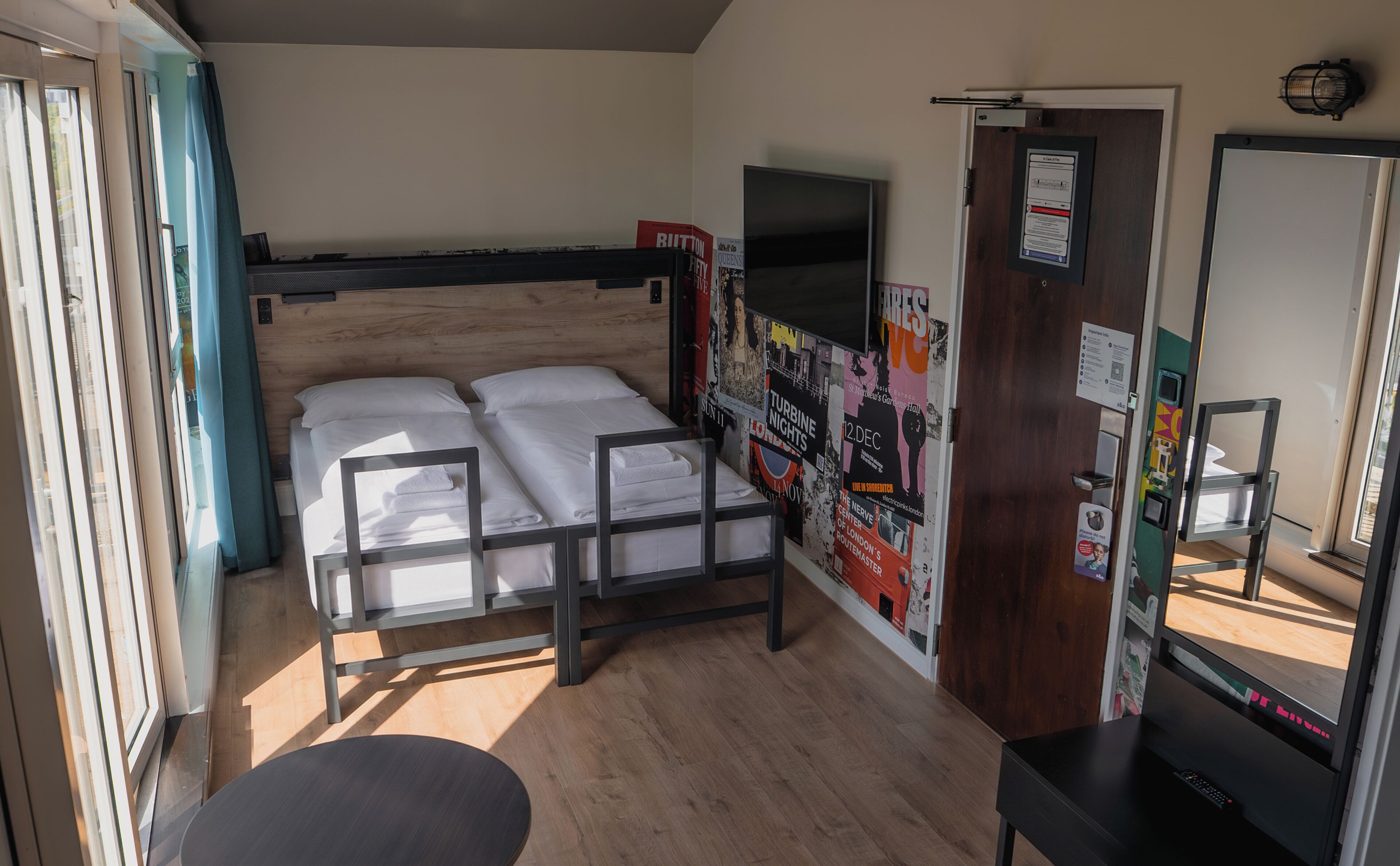 Una cama individual con cabecera, una mesita pequeña, un televisor montado en la pared y una puerta con un cartel.