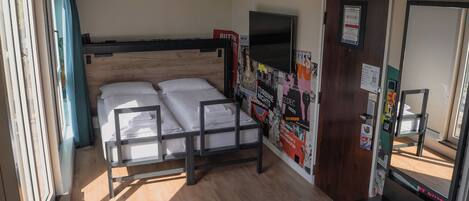 Una cama individual con cabecera, una mesita pequeña, un televisor montado en la pared y una puerta con un cartel.
