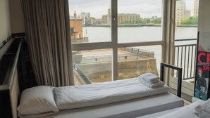 A hotel room with a bed, a piano, a large window with a view of a river and buildings.