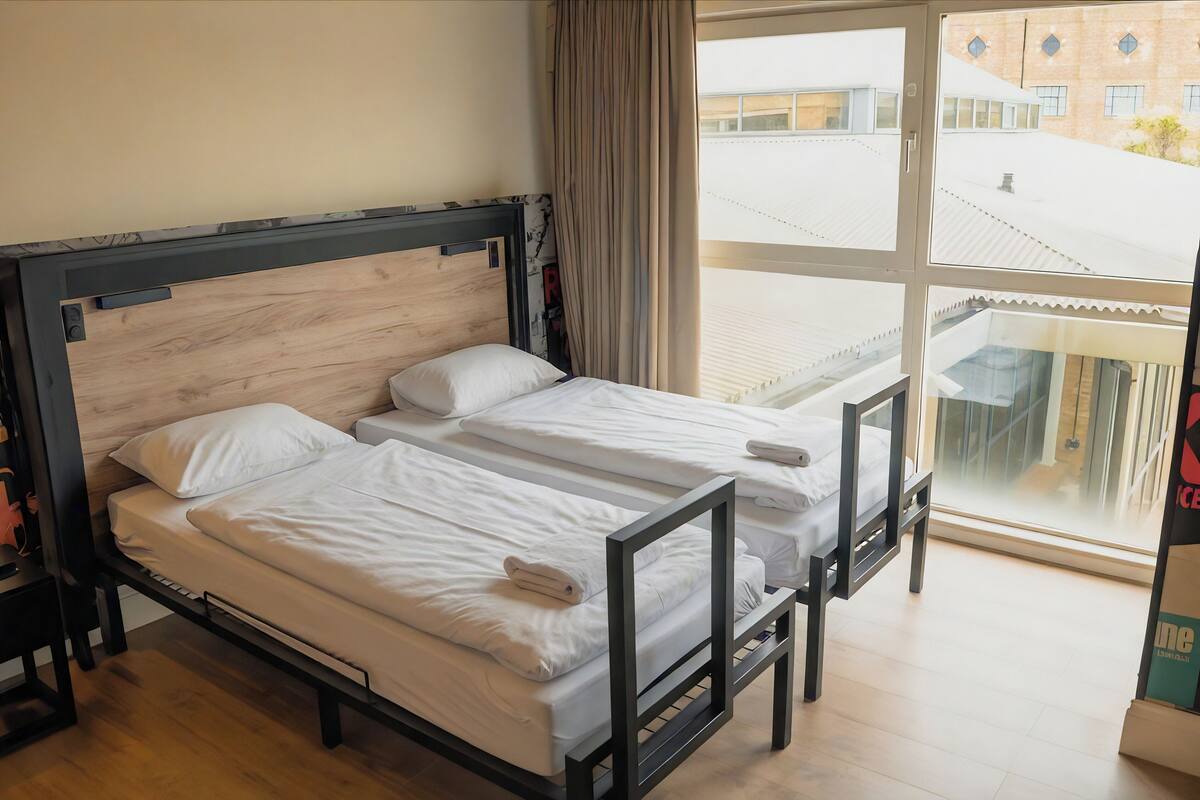 A room with two beds, a wooden headboard, a window with curtains, and a view of buildings outside.
