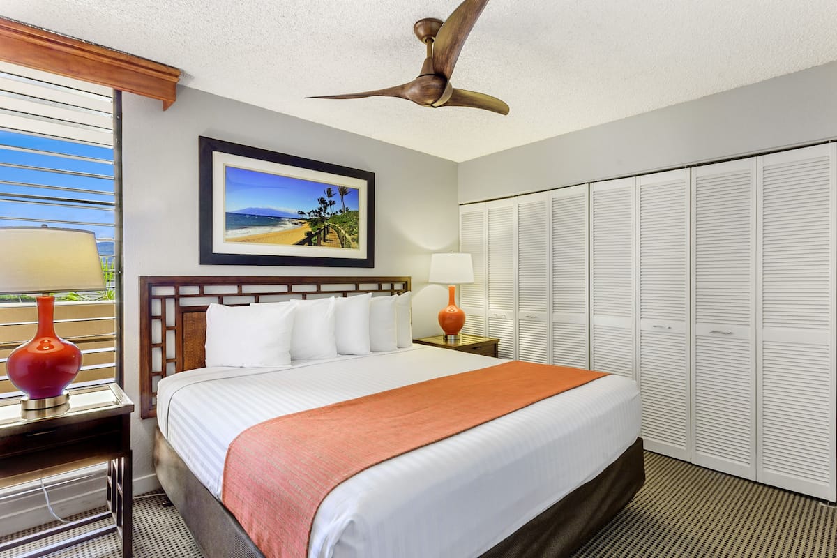 A hotel room with a bed, a ceiling fan, a nightstand with a lamp, a framed picture on the wall, and a closet with louvered doors.